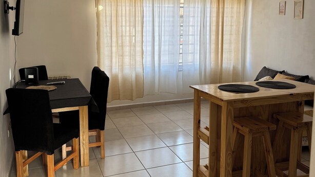 Un coin repas avec une table et des chaises noires, une table en bois avec des tabourets, un luminaire au plafond et une fenêtre ornée de rideaux légers.