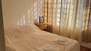 Une chambre avec un couvre-lit blanc et moelleux, une table de chevet en bois avec une lampe, et des rideaux légers qui laissent filtrer la lumière du soleil.