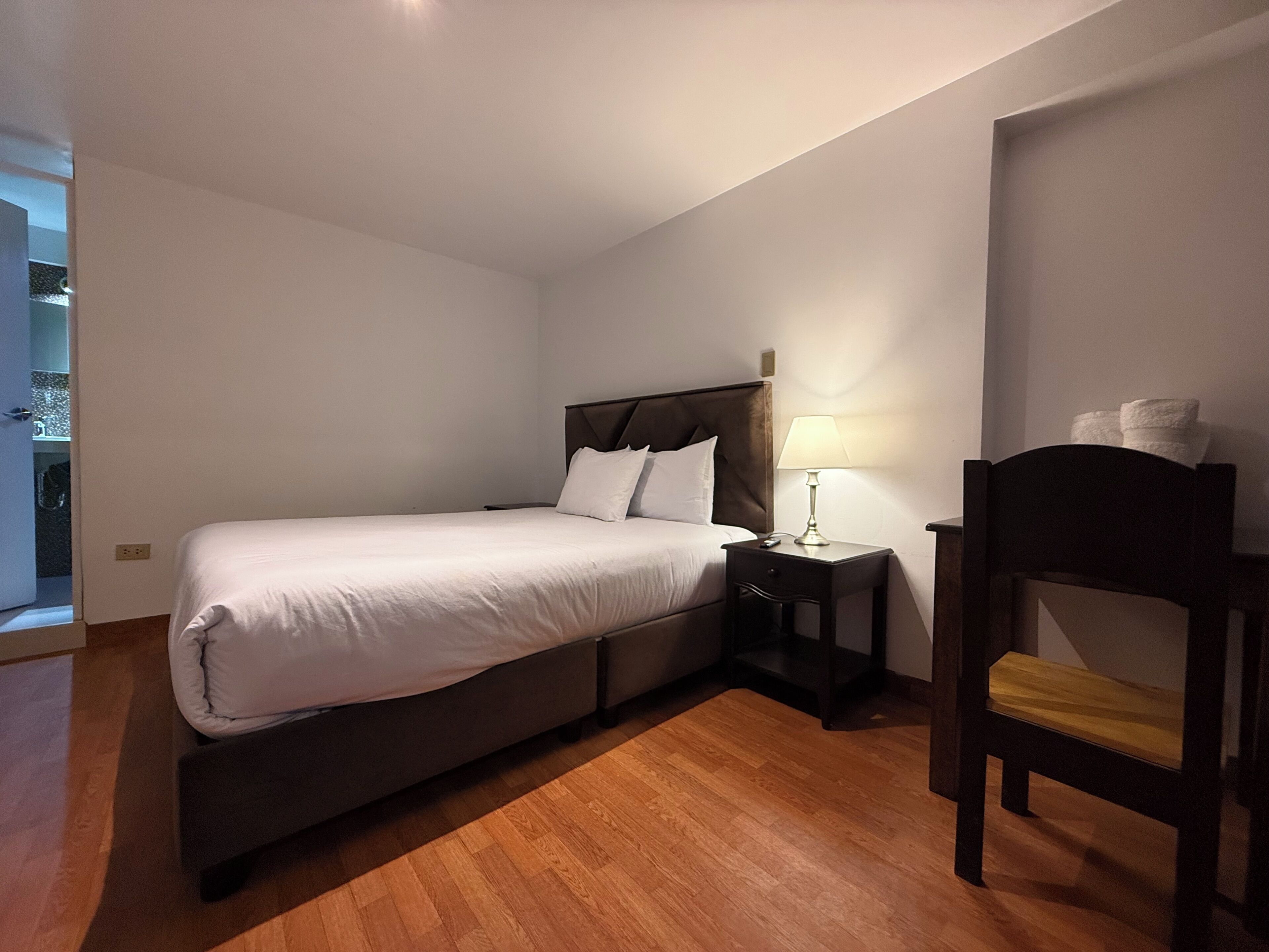 A bedroom with a bed, bedside table, lamp, chair, and a view of a bathroom.
