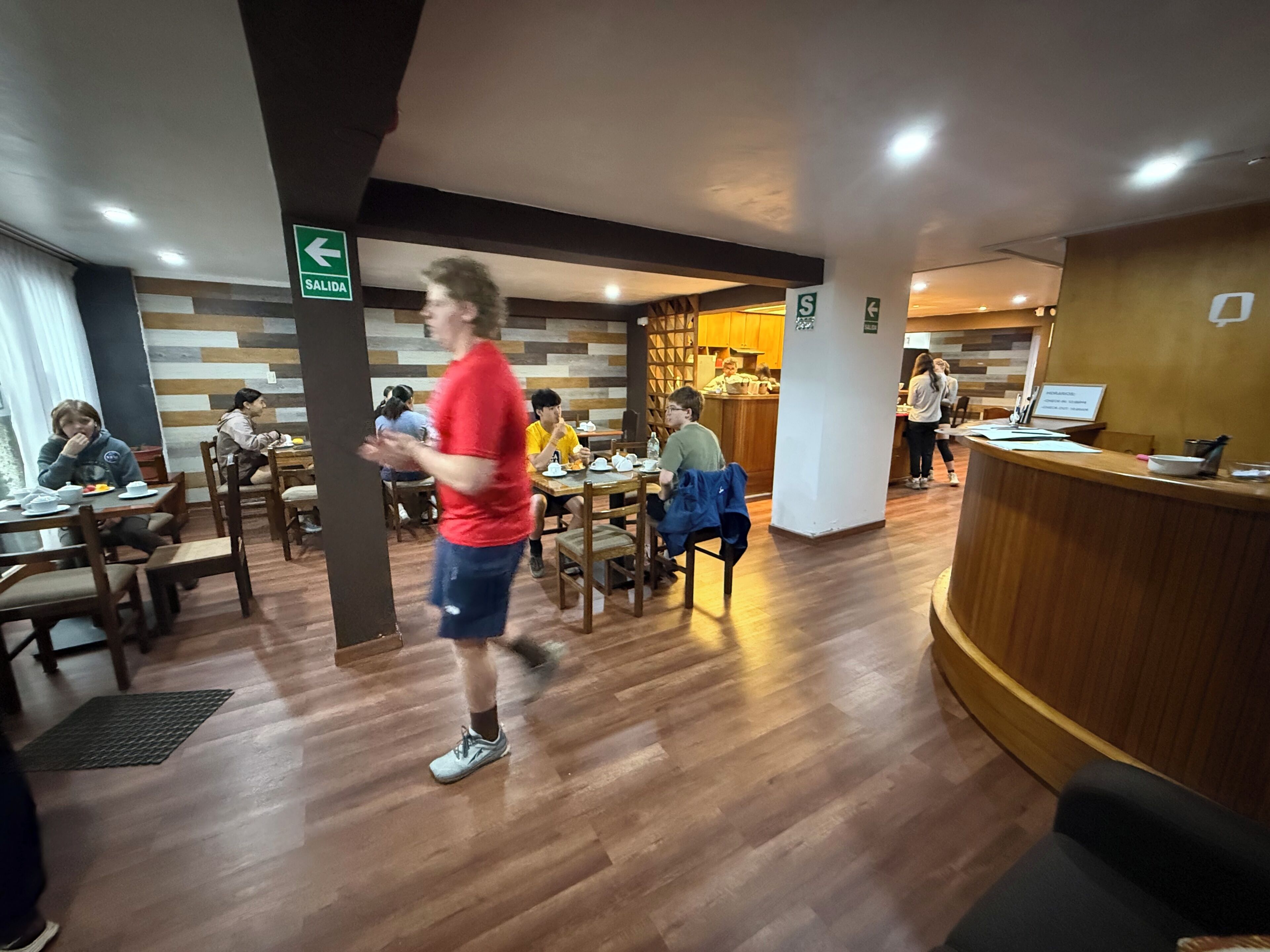 Un restaurante con suelo de madera, mesas, sillas y una barra. Se ven señales que indican una salida y un baño.