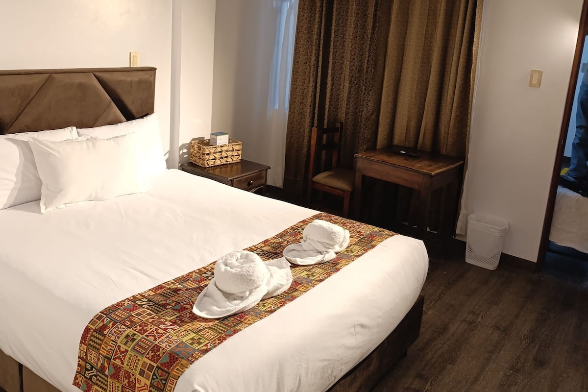 A hotel room with a large bed, a wooden headboard, a small desk, a chair, and a window with curtains.