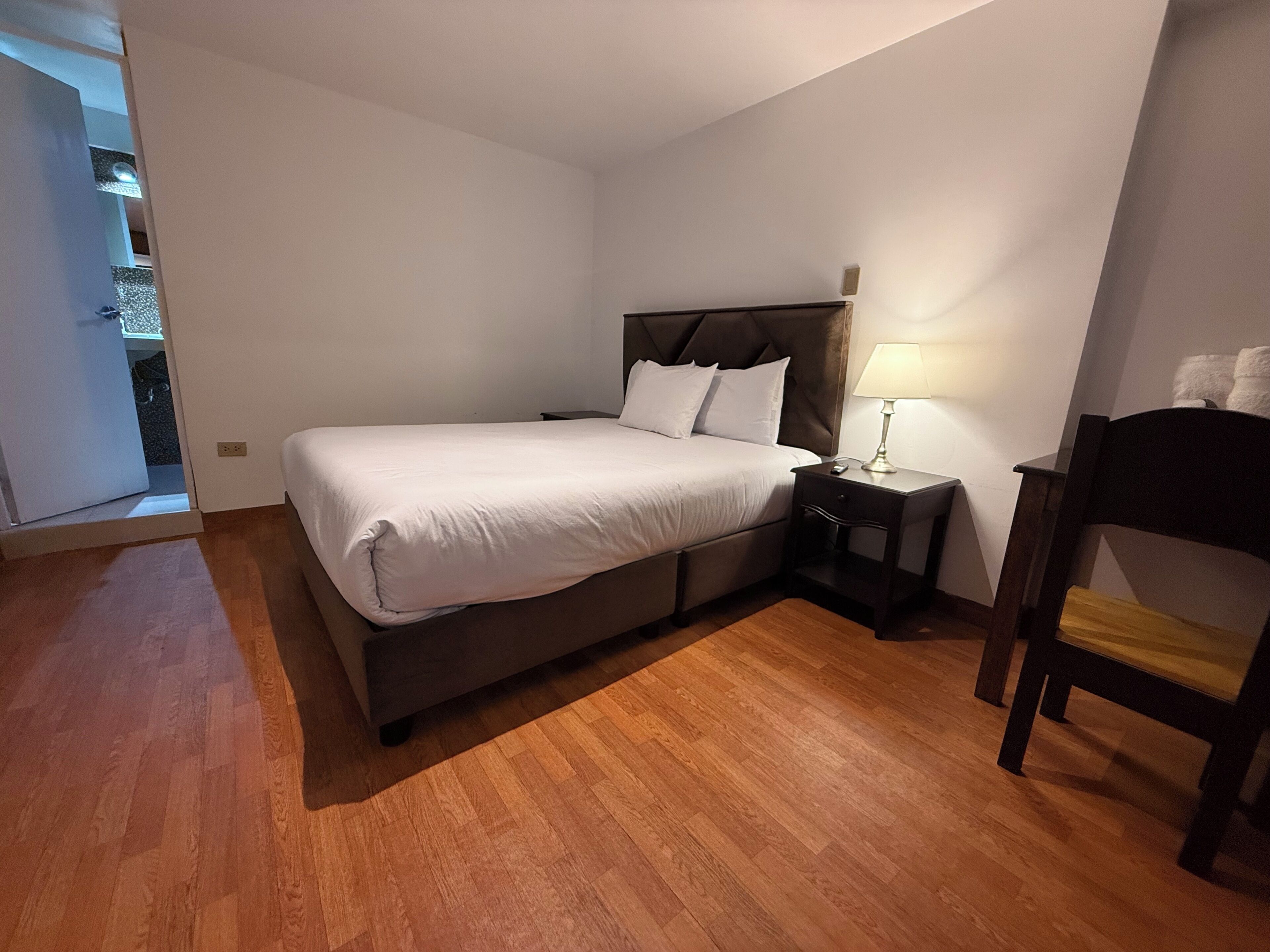 A bedroom with a bed, bedside table, lamp, chair, and wooden flooring.