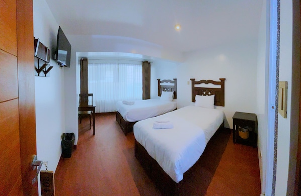 Habitación de hotel con dos camas individuales, un escritorio, una silla, un televisor montado en la pared y una mesita con un cubo de basura.