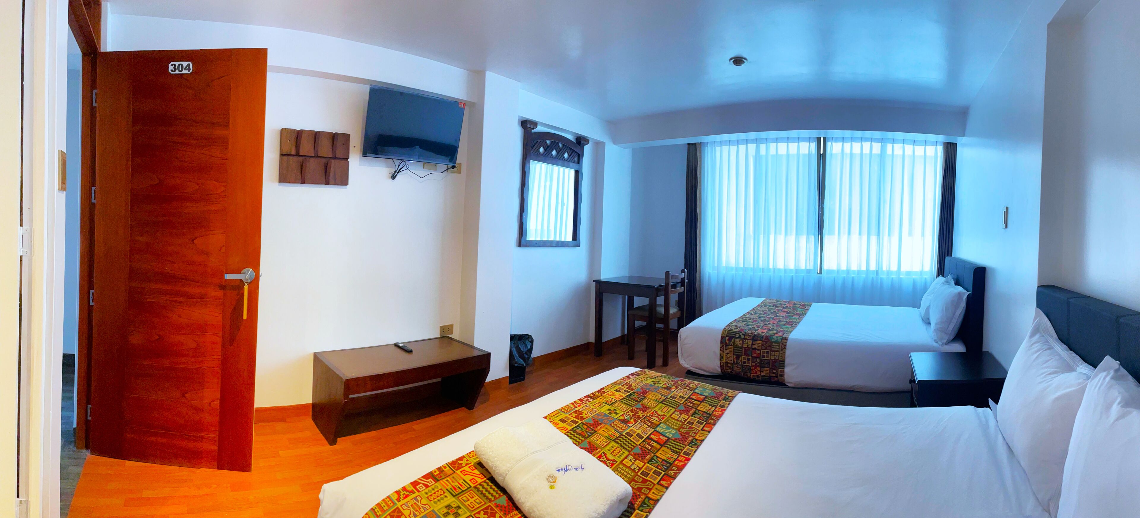 A hotel room with two beds, a TV mounted on the wall, a desk, and a window with curtains.