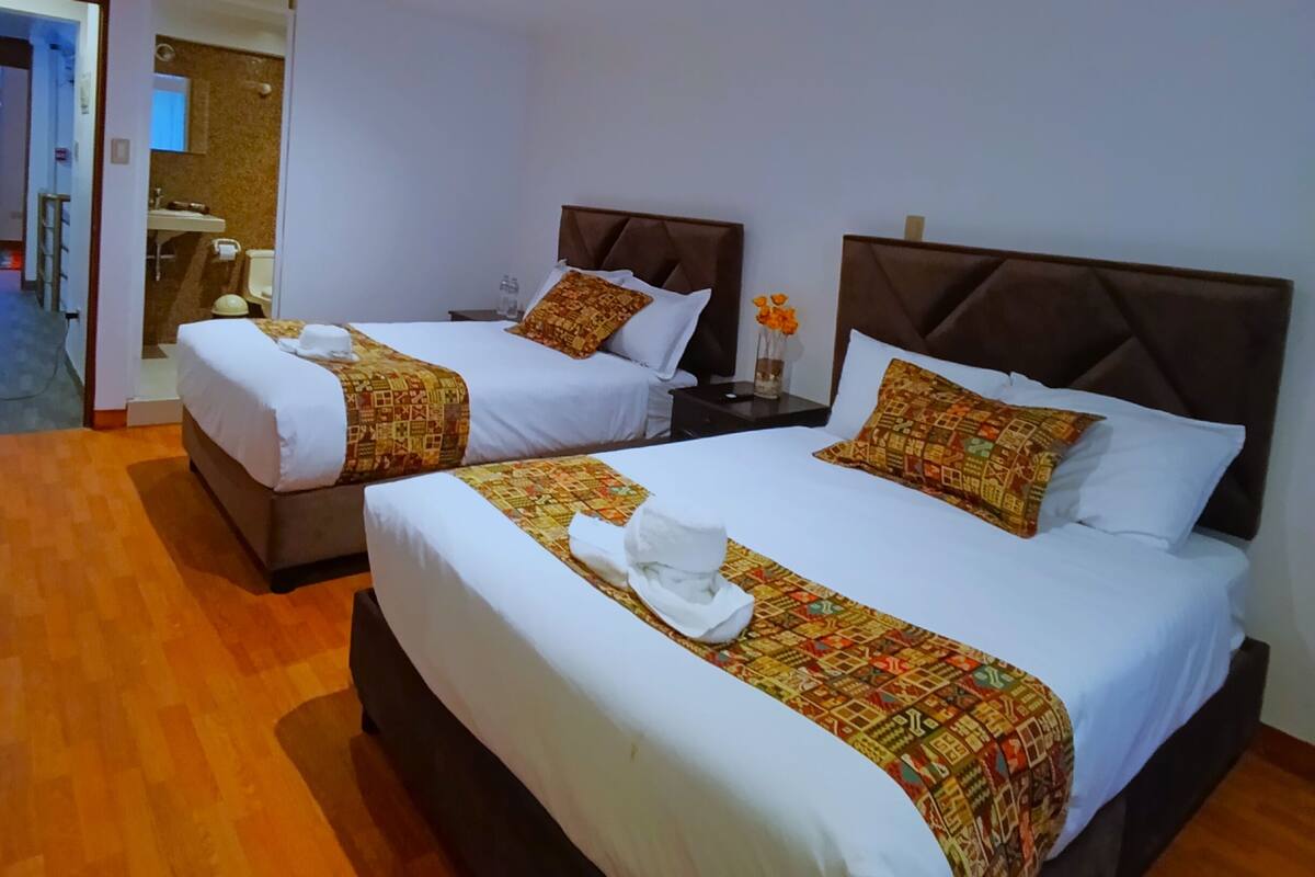 A hotel room with two beds, each with a patterned bedspread and pillows, a wooden floor, and a bathroom visible in the background.