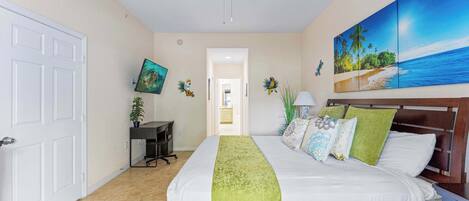 Un dormitorio con cama, ventilador de techo, escritorio, silla, un cuadro y vista a la playa.