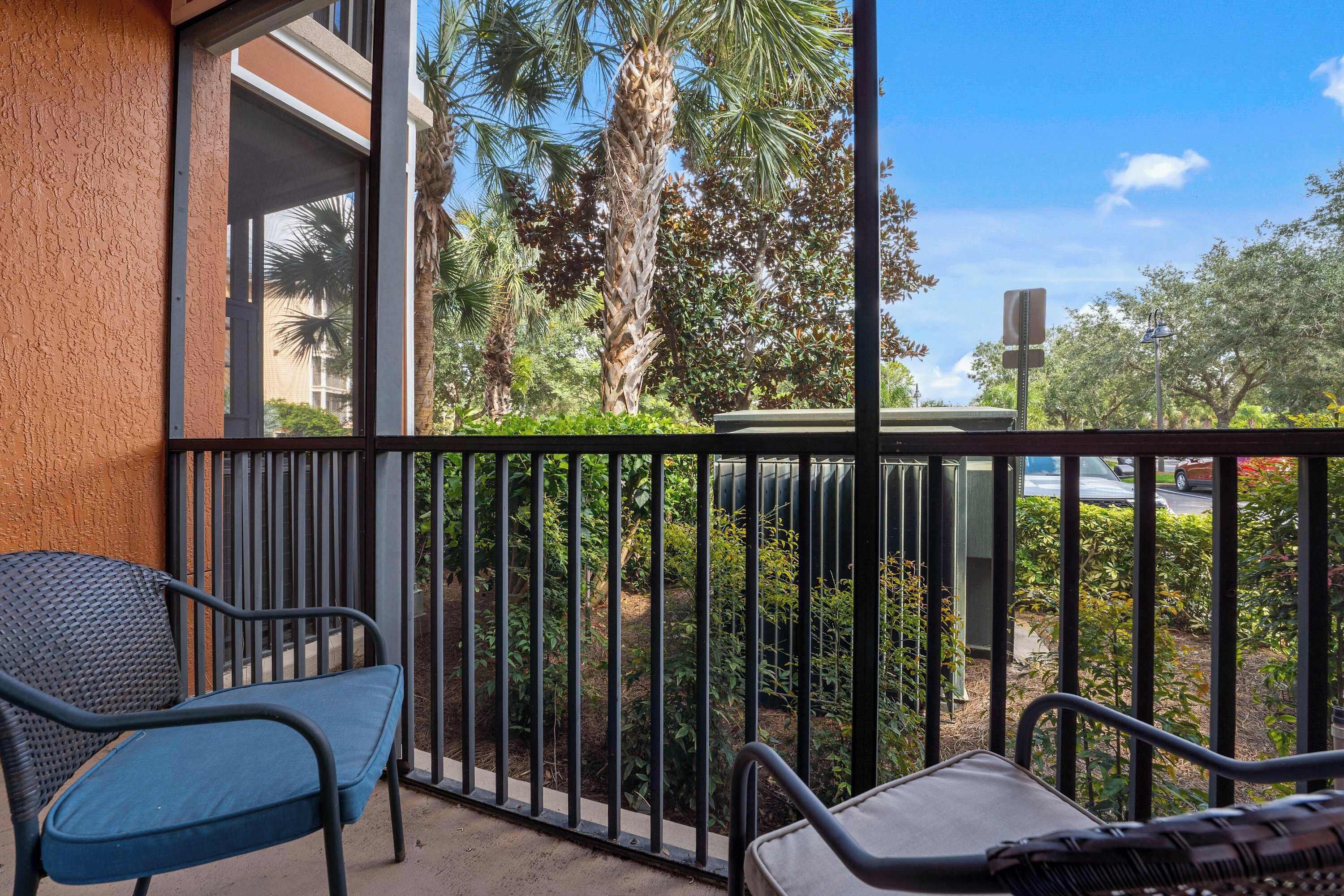 A balcony with two chairs, a view of a garden with palm trees, and a building in the background.