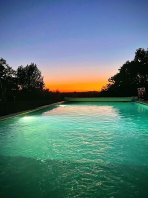Ein Schwimmbad mit klarem Wasser, umgeben von Bäumen und einem Sonnenuntergang im Hintergrund.