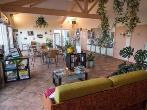 A spacious living area with a sofa, chairs, a coffee table, and various plants. There is a bookshelf with books and a small table with a vase. The room has a tiled floor and a wooden ceiling.