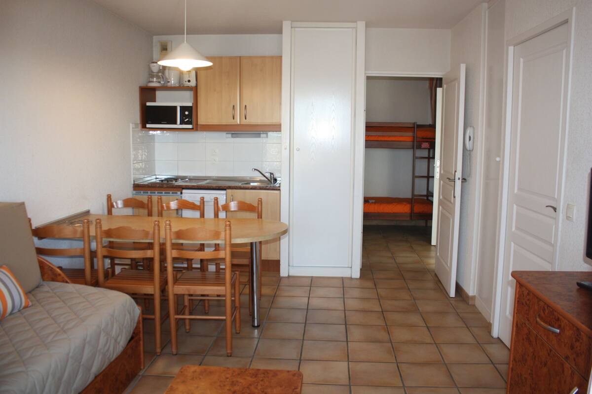 Una habitación compacta con una cama, una mesa de comedor con sillas, una kitchenette con microondas y una puerta que da a una zona con literas.