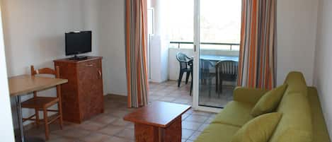 Ein kompakter Wohnraum mit Sofa, Fernseher auf einem Schrank, Esstisch und Balkon mit einem Stuhl.
