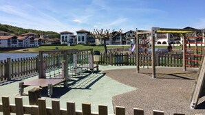 A playground with climbing structures and a fenced area, set against a backdrop of residential buildings and a body of water.