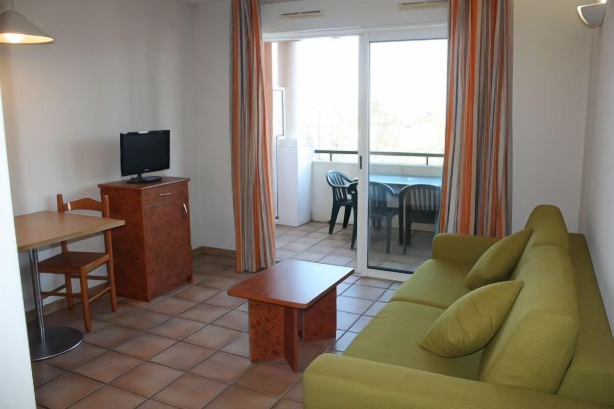 A living room with a sofa, a TV on a wooden cabinet, a table, and a balcony with chairs.