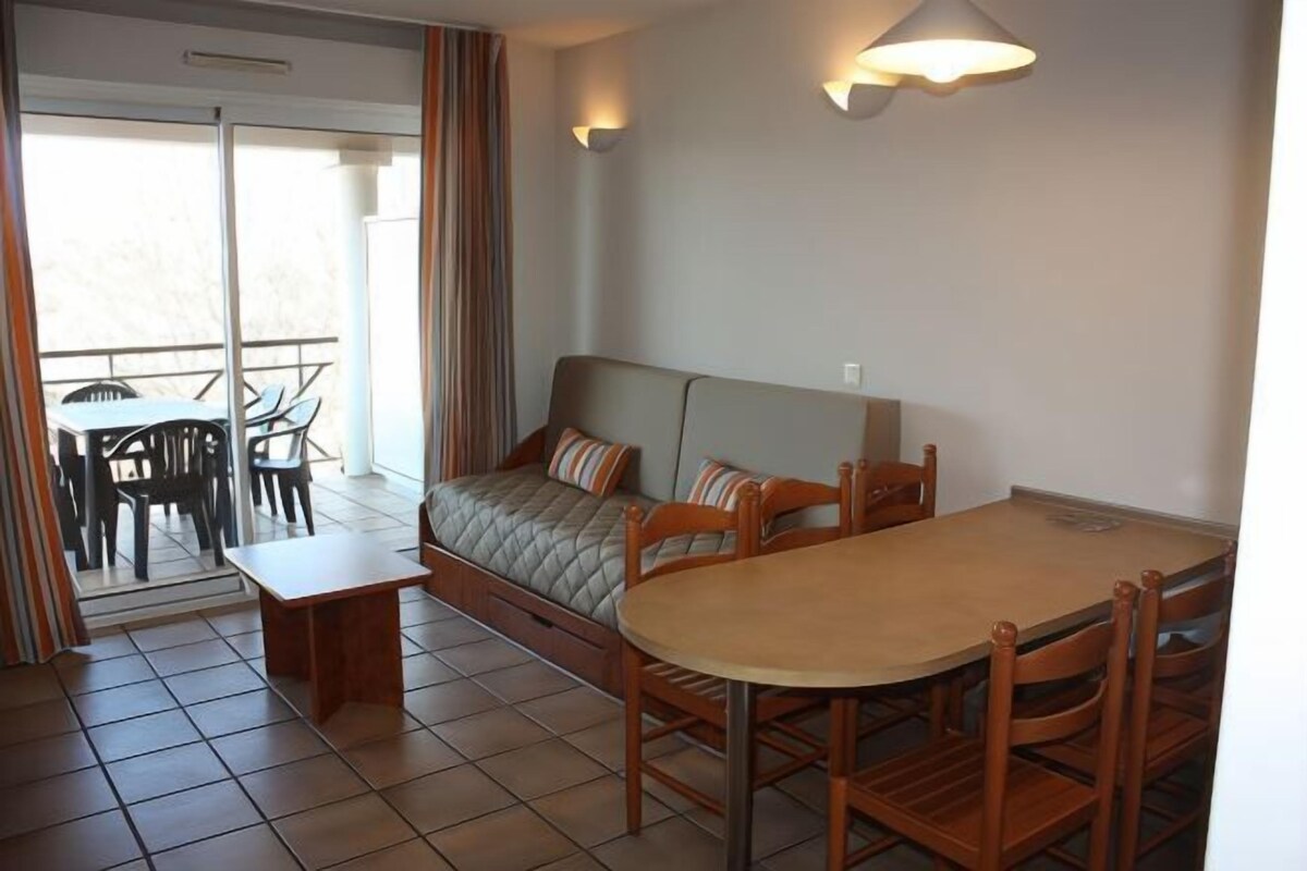 A room with a sofa bed, a dining table with chairs, a small table, and a balcony with chairs.