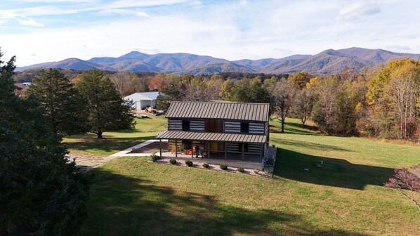 A log cabin with a porch, surrounded by a grassy yard and trees, with mountains in the background.