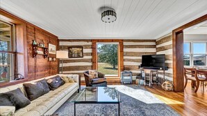 A log cabin interior with a sofa, coffee table, TV, and a large window offering a view of the outdoors. - Charming, fully renovated 19th century cabin with spectacular views. 4 BR, 2 BA. (Arrington)