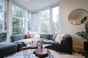 Een moderne woonkamer met een grijze modulaire bank, een ronde spiegel en een groot raam met uitzicht op groen.