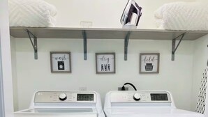 A laundry room with two white front-loading washing machines and dryers, a shelf with towels and a laundry basket, and three framed signs on the wall reading "wash," "dry," and "fold." - Modern 2-bedroom house, walking distance to beach, restaurants and shopping. (Miramar Beach)