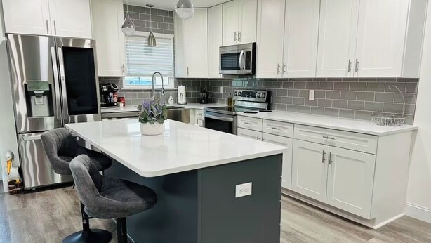 A modern kitchen with white cabinets, stainless steel appliances, a central island with bar stools, and a grey backsplash. - Modern 2-bedroom house, walking distance to beach, restaurants and shopping. (Miramar Beach)