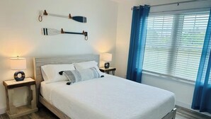 A neatly made bed with a white comforter and pillows, two bedside lamps, a wooden nightstand, a wall-mounted paddleboard, and a window with blue curtains. - Modern 2-bedroom house, walking distance to beach, restaurants and shopping. (Miramar Beach)