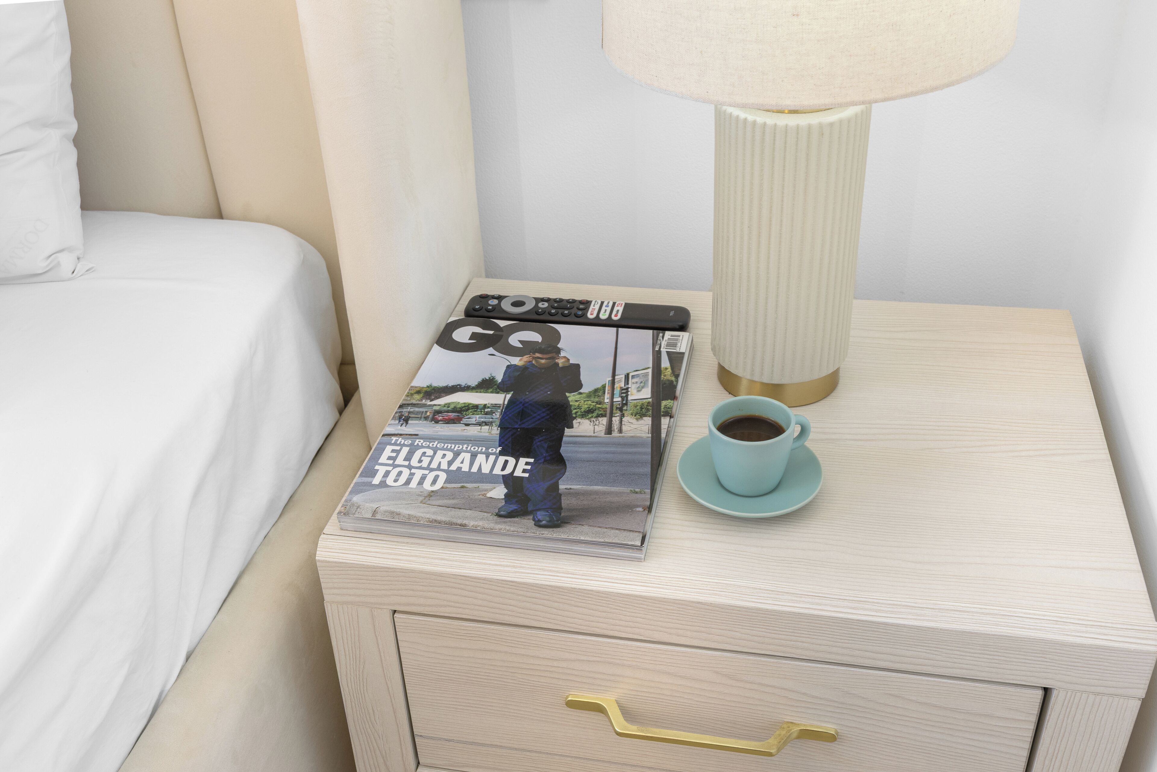 Un llit ben fet, una revista amb un home a la portada, un comandament a distància, una làmpada i una tassa de cafè sobre una taula de nit.