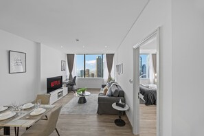A modern living room with a flat-screen TV, a dining area with a table set for four, a grey sofa, and a city view through large windows. - Your Stay Corporate Residences DIFC (Dubai)