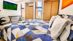 Un dormitorio con una cama grande, un televisor instalado en la pared, un armario y una ventana con vista a edificios.