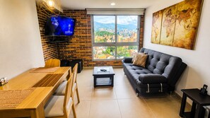 Ein Wohnzimmer mit einer Couch, einem Couchtisch, einem an einer Ziegelwand montierten Fernseher und einem großen Fenster mit Blick auf die Stadt.