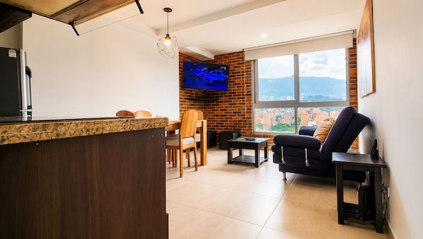 A modern living area with a kitchen counter, dining table, chairs, a sofa, a TV mounted on a brick wall, and a large window offering a view.