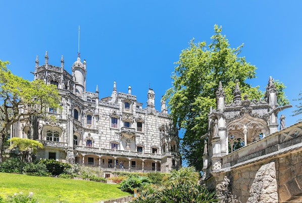 A grand historic building with multiple towers and ornate architecture, surrounded by lush greenery and a stone bridge.