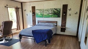 Un dormitorio con cama, una silla, ventana con cortinas, un cuadro en la pared y suelo de madera.