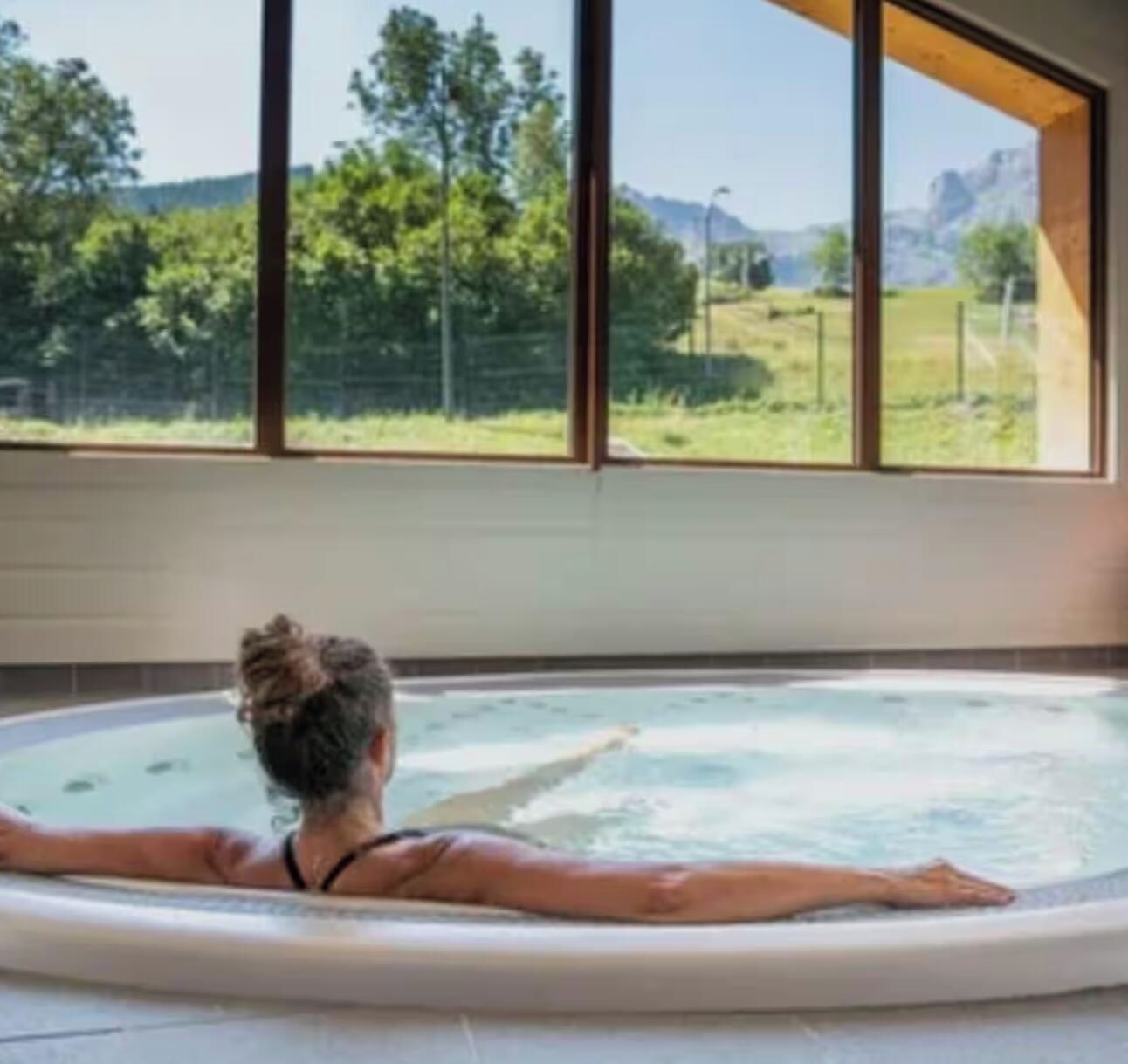 A person relaxing in a hot tub with a view of a mountainous landscape through large windows.