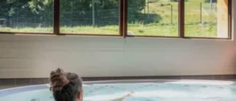 Une personne se détend dans un bain à remous, avec vue sur un paysage montagneux grâce à de grandes fenêtres.