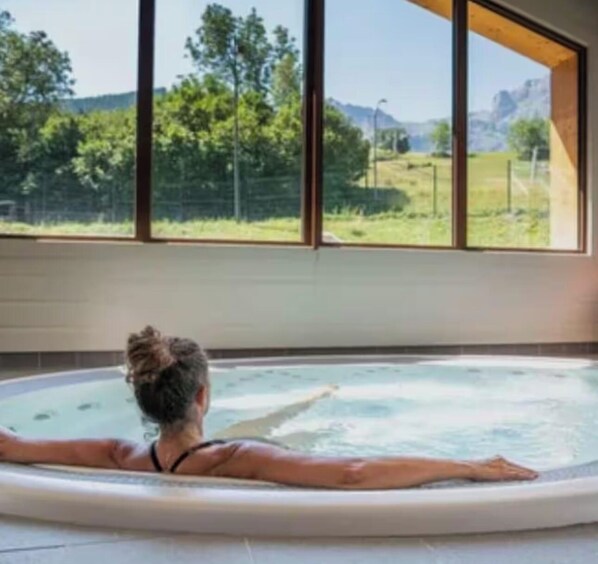 A person relaxing in a hot tub with a view of a green landscape through large windows.