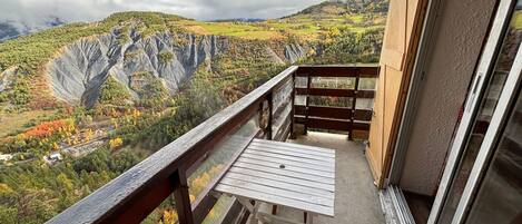 A balcony with a table and chairs, overlooking a mountainous landscape with autumn foliage.