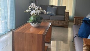 En moderne stue med et træ sofabord, en grå sofa og en fletstol. Der er et billede på væggen og et vindue med lette gardiner.
