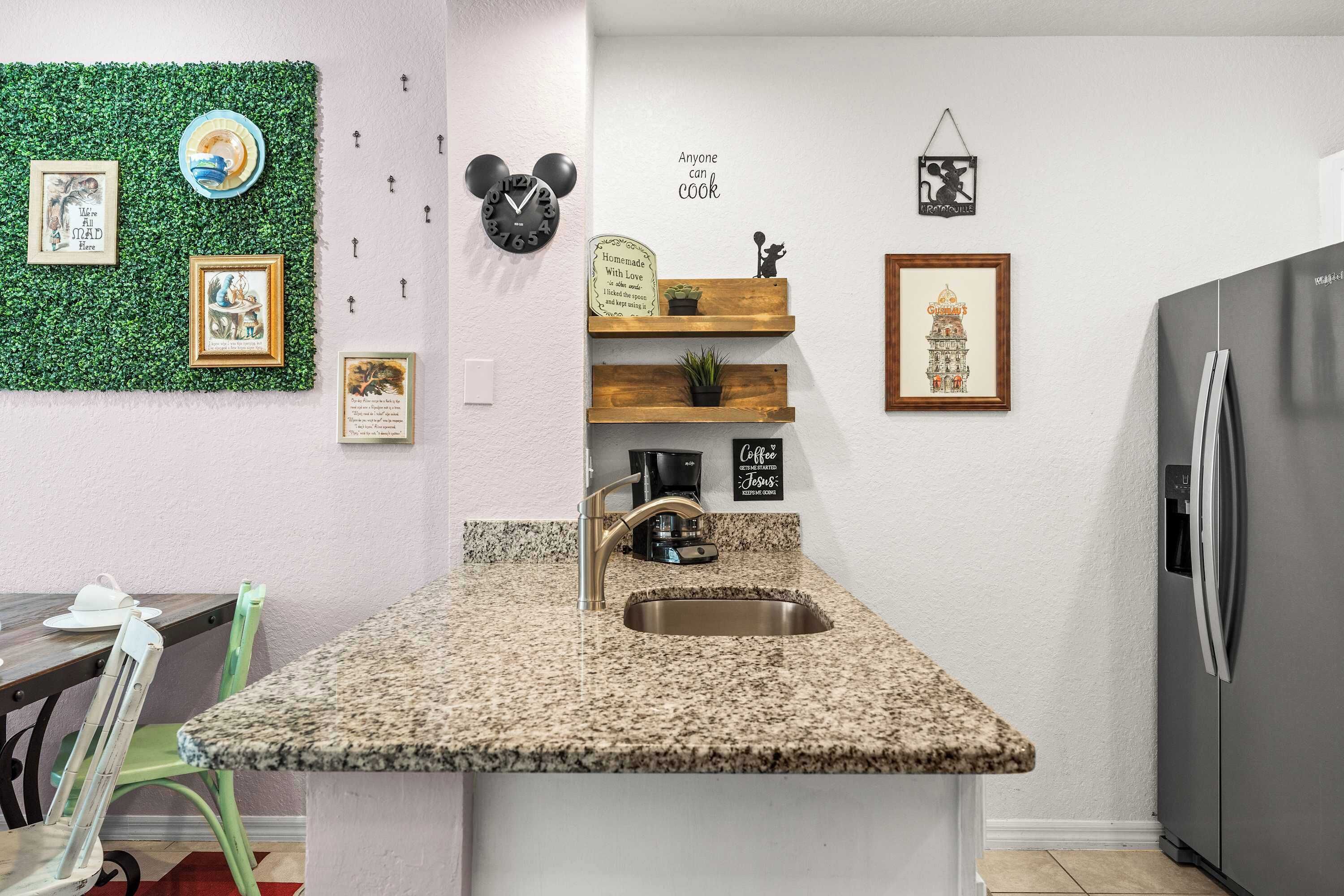 Cozinha com bancada de granito, pia de aço inoxidável e geladeira. Prateleiras fixadas na parede exibem objetos decorativos e um relógio.