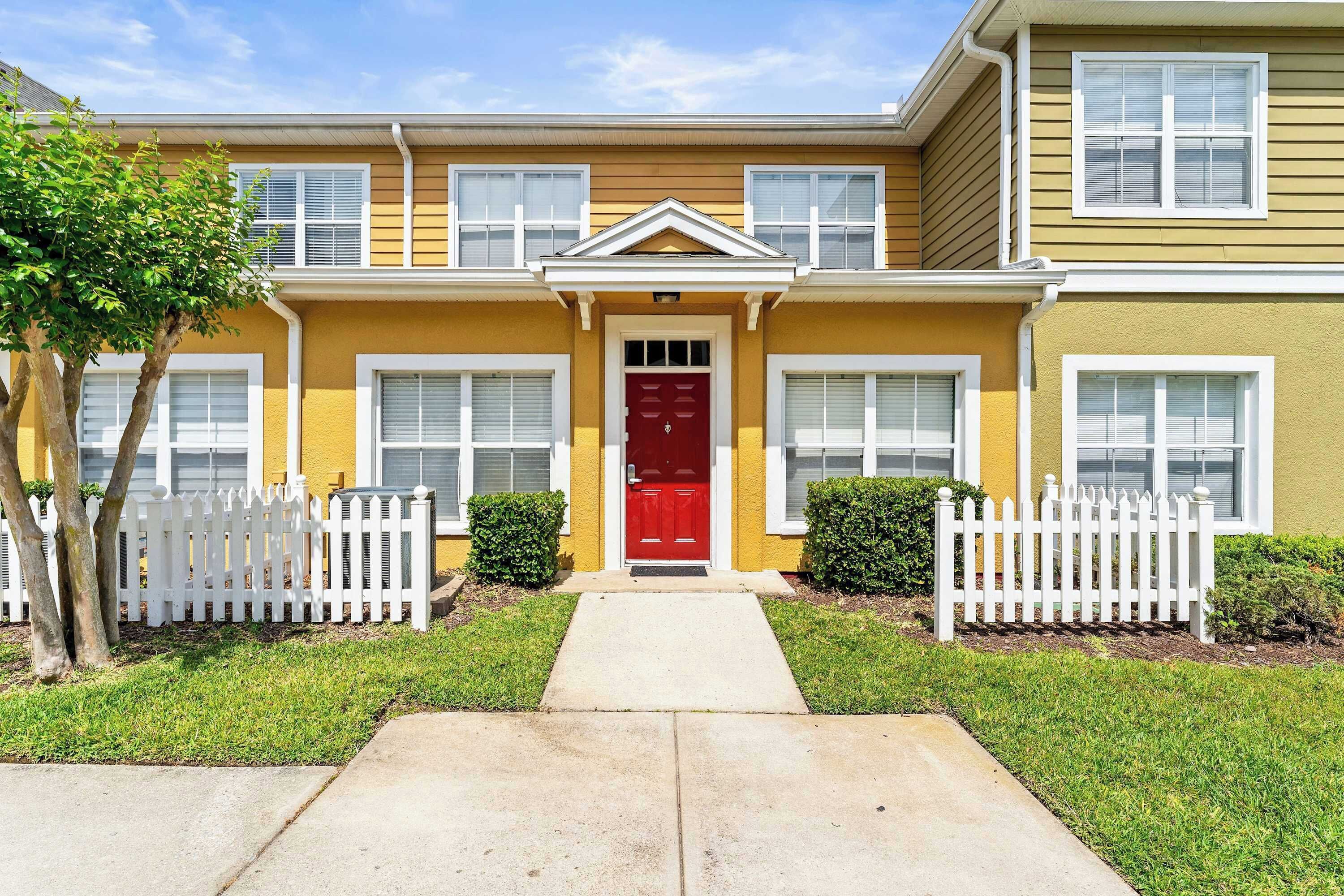 A two-story residential building with a red front door, white picket fence, and a small front yard.