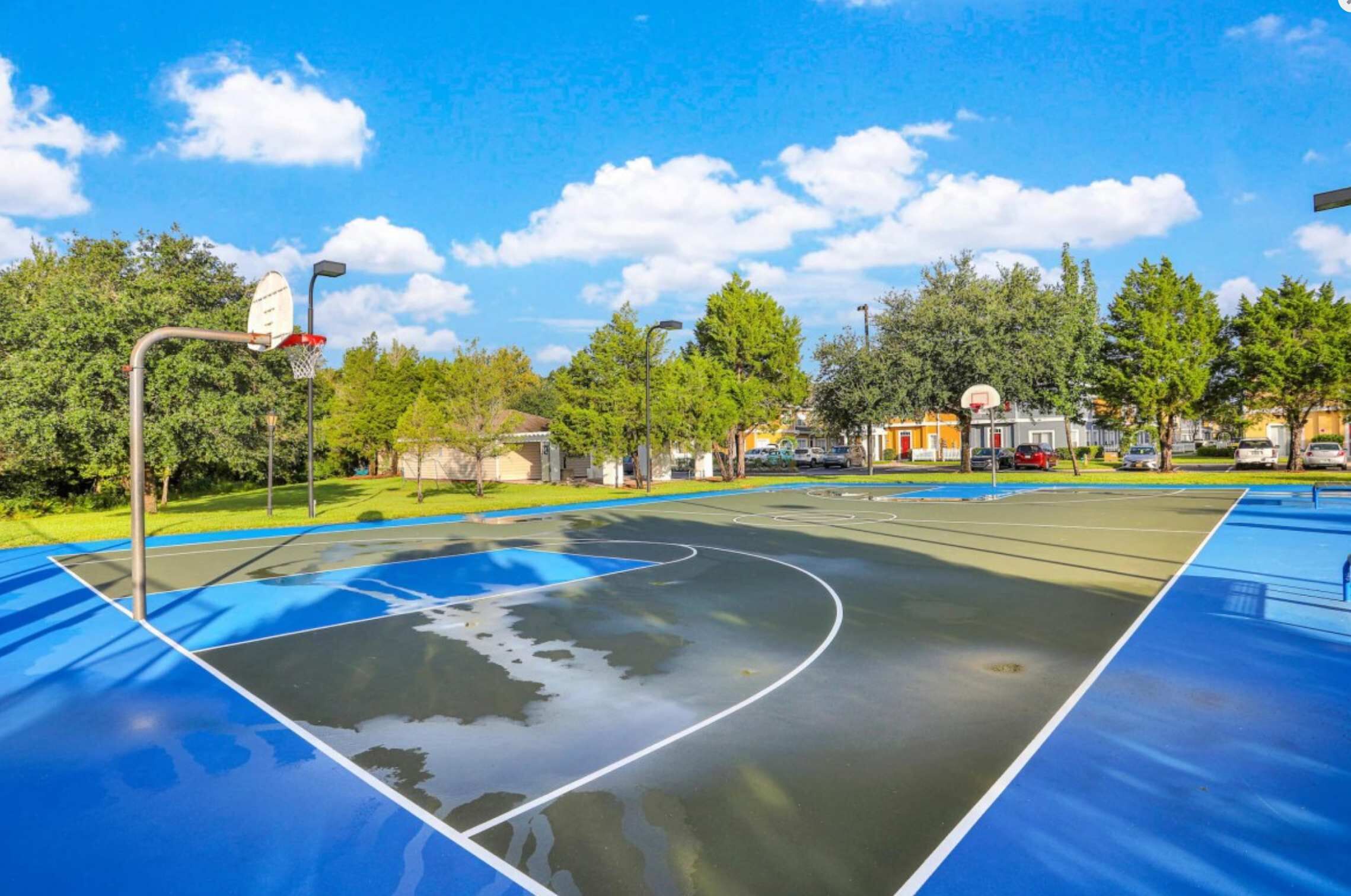 An outdoor basketball court with a blue surface, surrounded by trees and residential buildings.