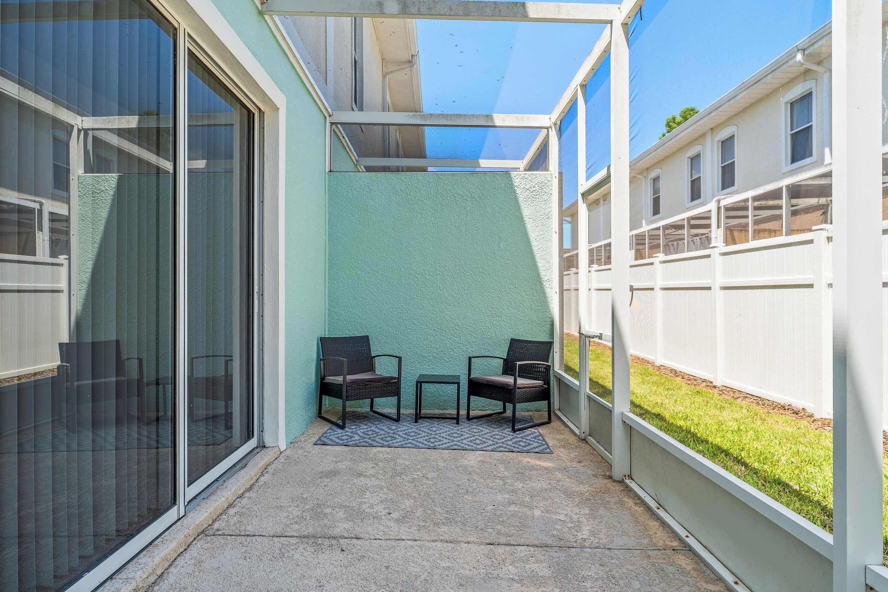 A covered patio area with two chairs and a small table, a sliding glass door, and a view of a white fence and greenery.