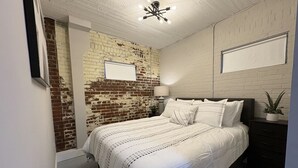 Een slaapkamer met een bakstenen muur, een wit bed, een nachtkastje en een plafondlamp.