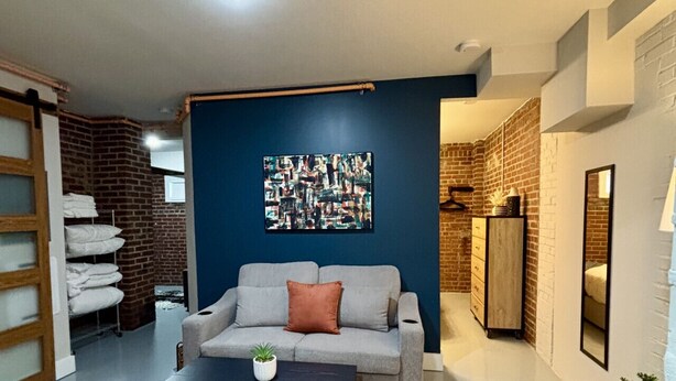 Una sala moderna con sofá, sillón y mesa de centro. Se observa un tapete de diseño geométrico, una obra de arte en la pared y una pared de ladrillo decorativa.