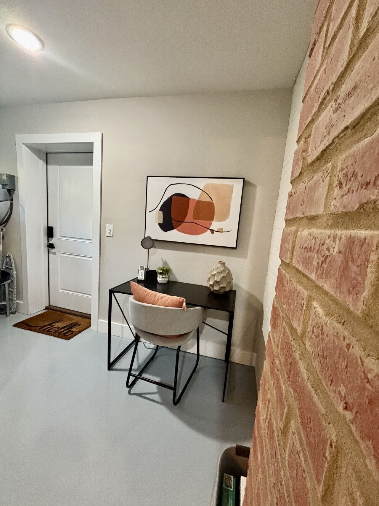 Un intérieur moderne avec un mur en briques, un bureau avec une chaise, un tableau abstrait encadré et une porte avec un paillasson.