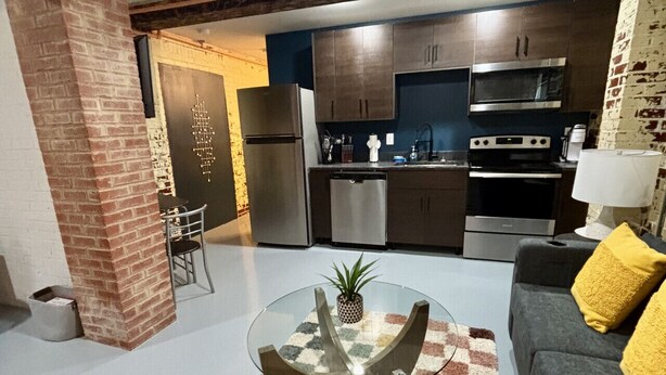Ein modernes Wohnzimmer mit einem Glastisch, einem Schachbrettteppich und einem grauen Sofa mit gelben Kissen. Im Hintergrund ist eine Küche mit dunklen Holzschränken und Edelstahlgeräten zu sehen.