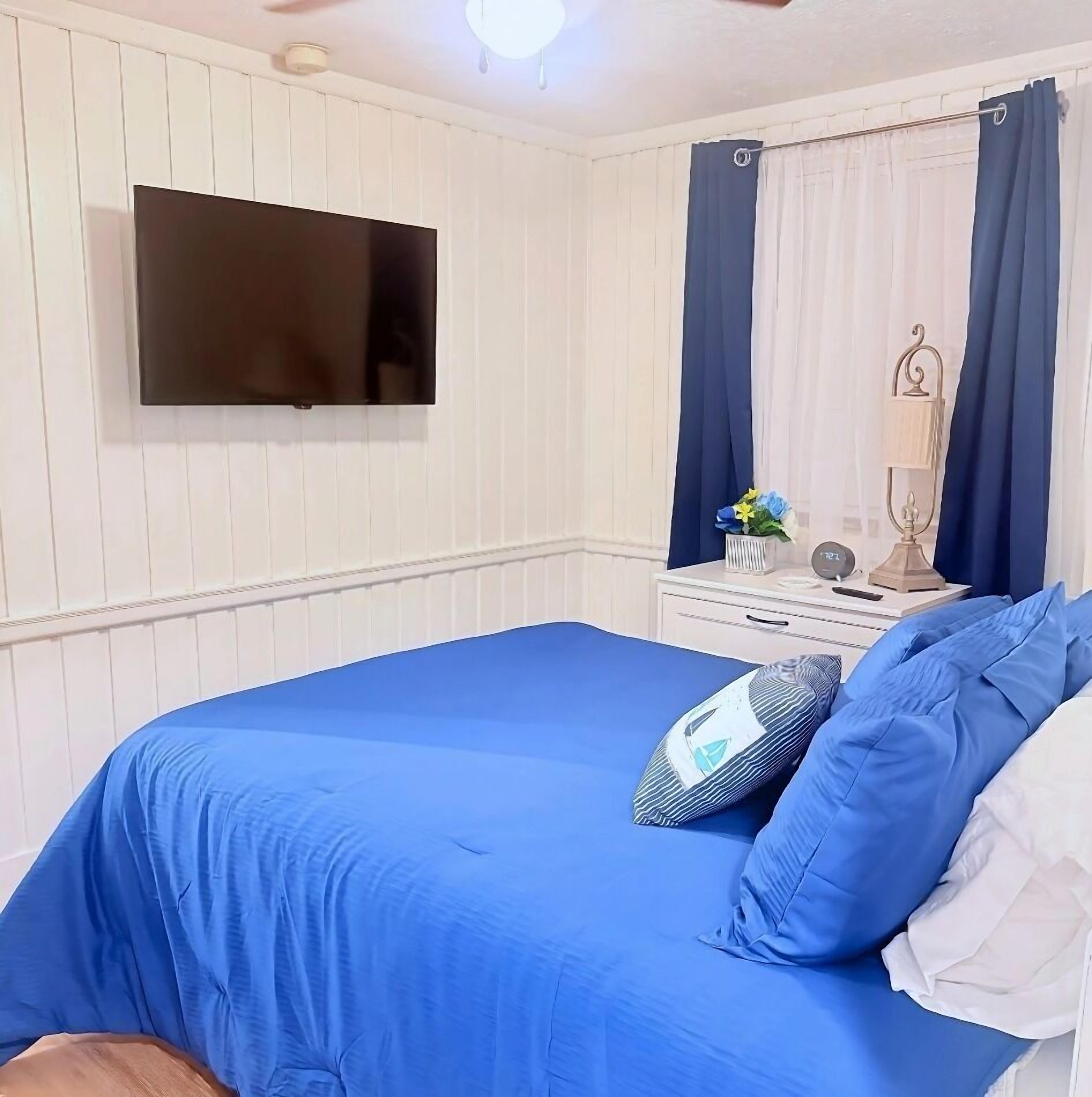 A bedroom with a bed, a TV mounted on the wall, a nightstand with a lamp, and a window with curtains.