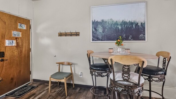 A dining area with a round wooden table, four chairs, a wall-mounted picture, and a door with a doormat.