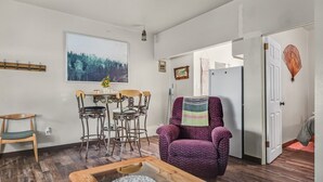 A living room with a wooden coffee table, a purple armchair, a dining table with chairs, and a large window with a forest view.