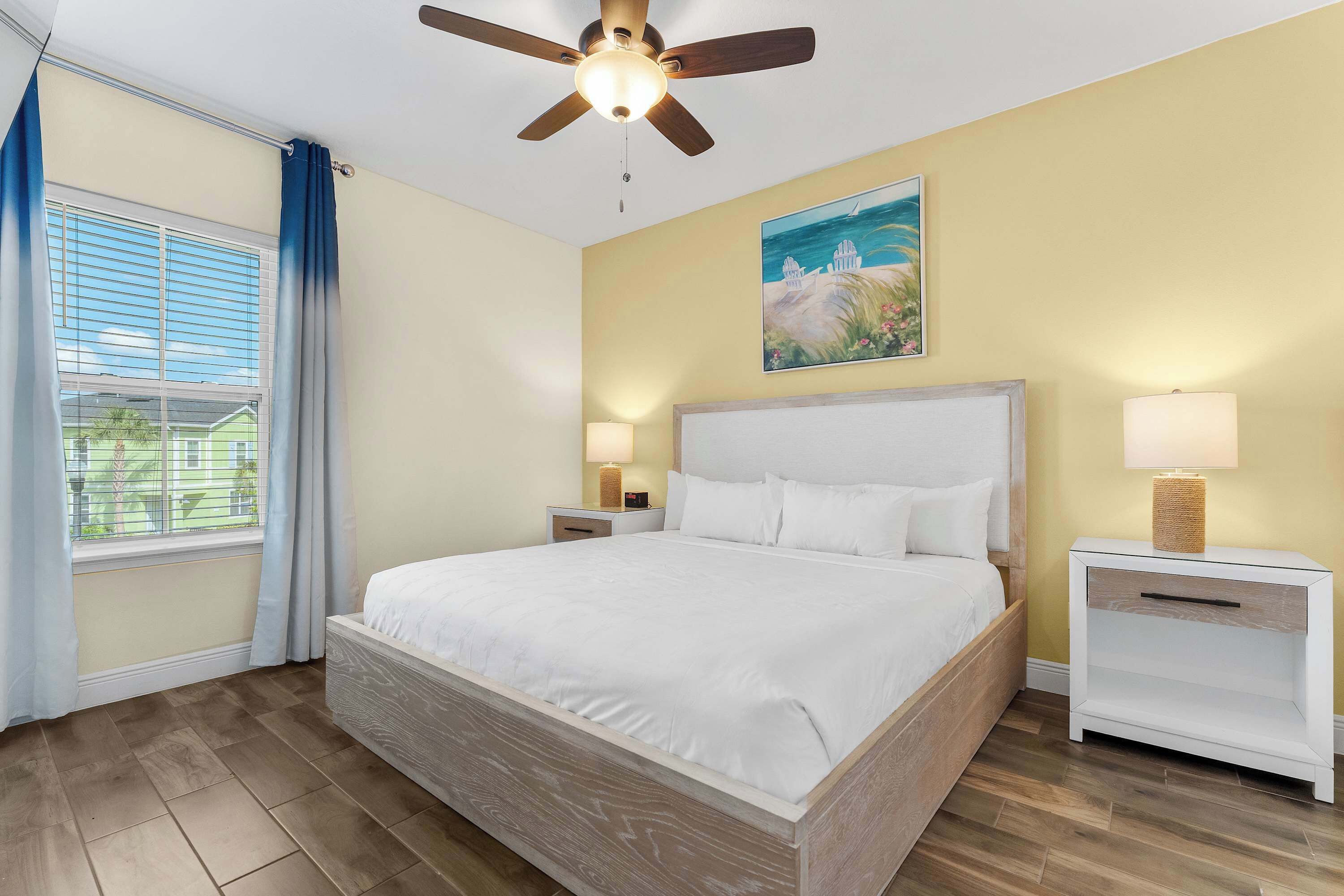 A bedroom with a large bed, two bedside tables with lamps, a ceiling fan, a window with curtains, and a painting on the wall.