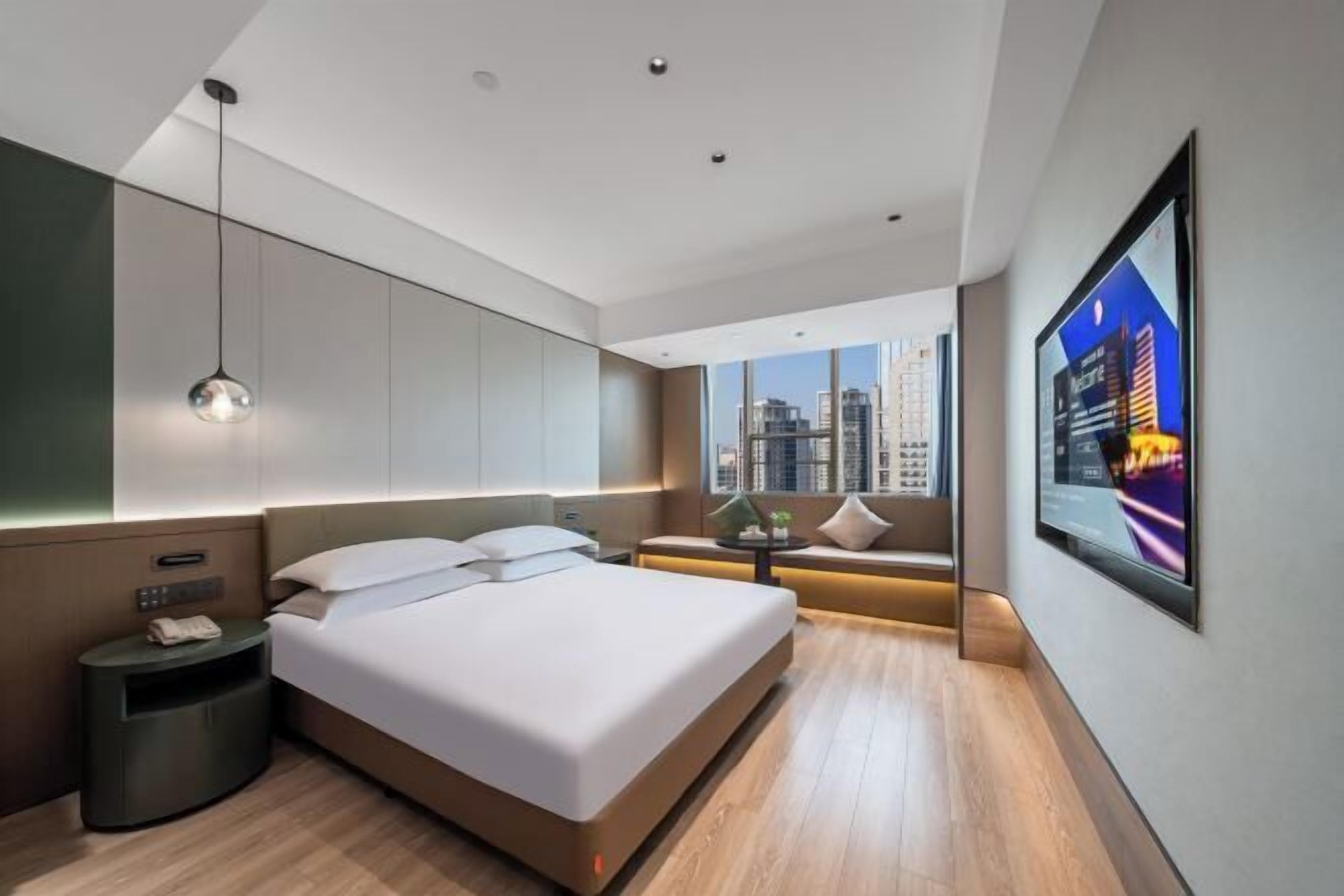 A modern hotel room with a large bed, a bedside table, a nightstand, a wall-mounted TV, and a view of a cityscape.