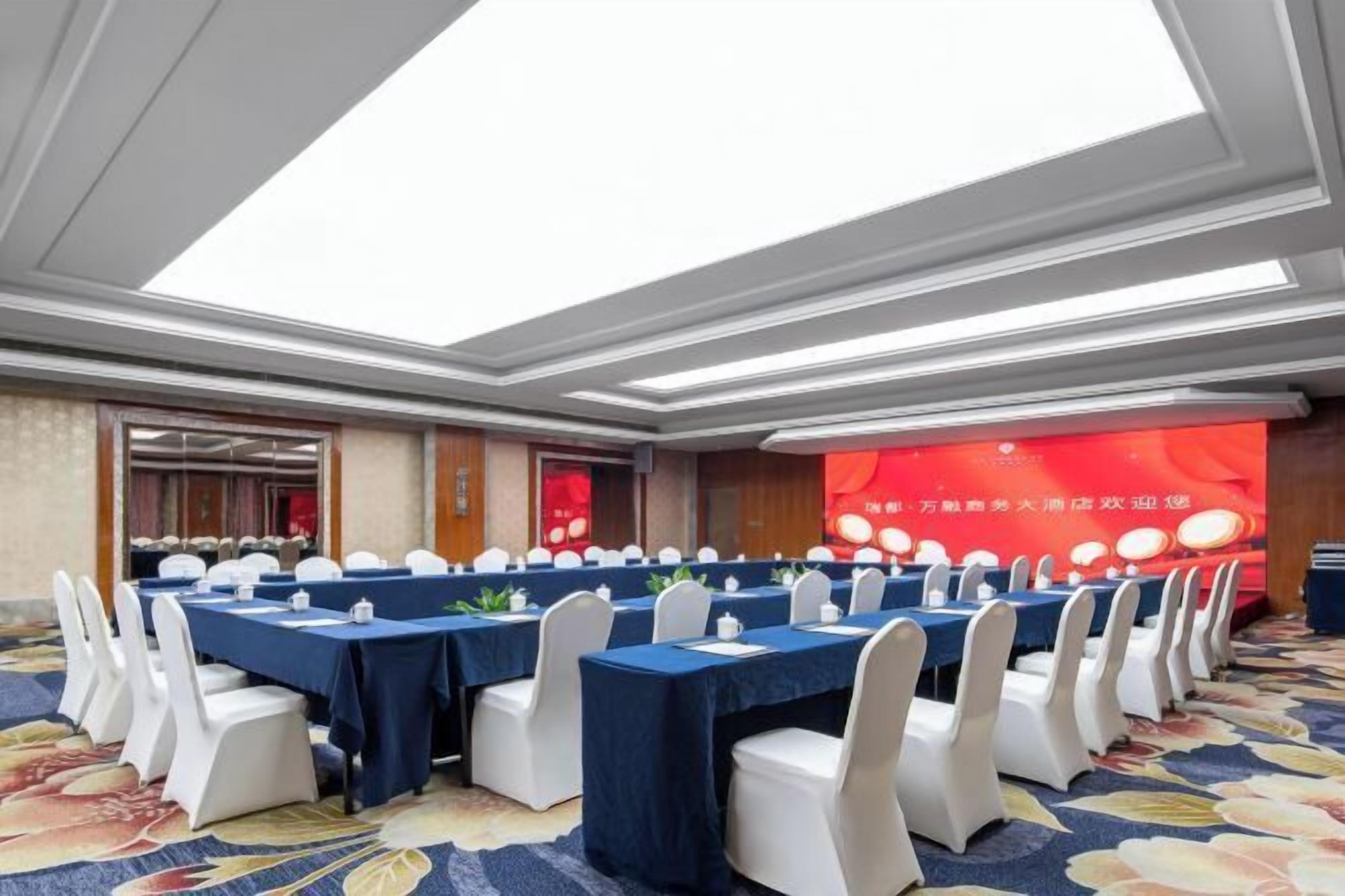 A conference room with multiple tables, white chairs, and a stage with a red backdrop.